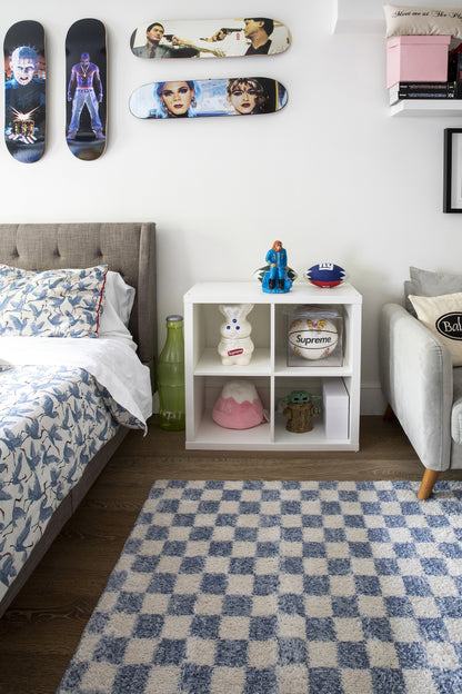 Kids' bedroom with white cube storage, blue check rug, gray upholstered bed, skateboard wall art, and playful figurines.