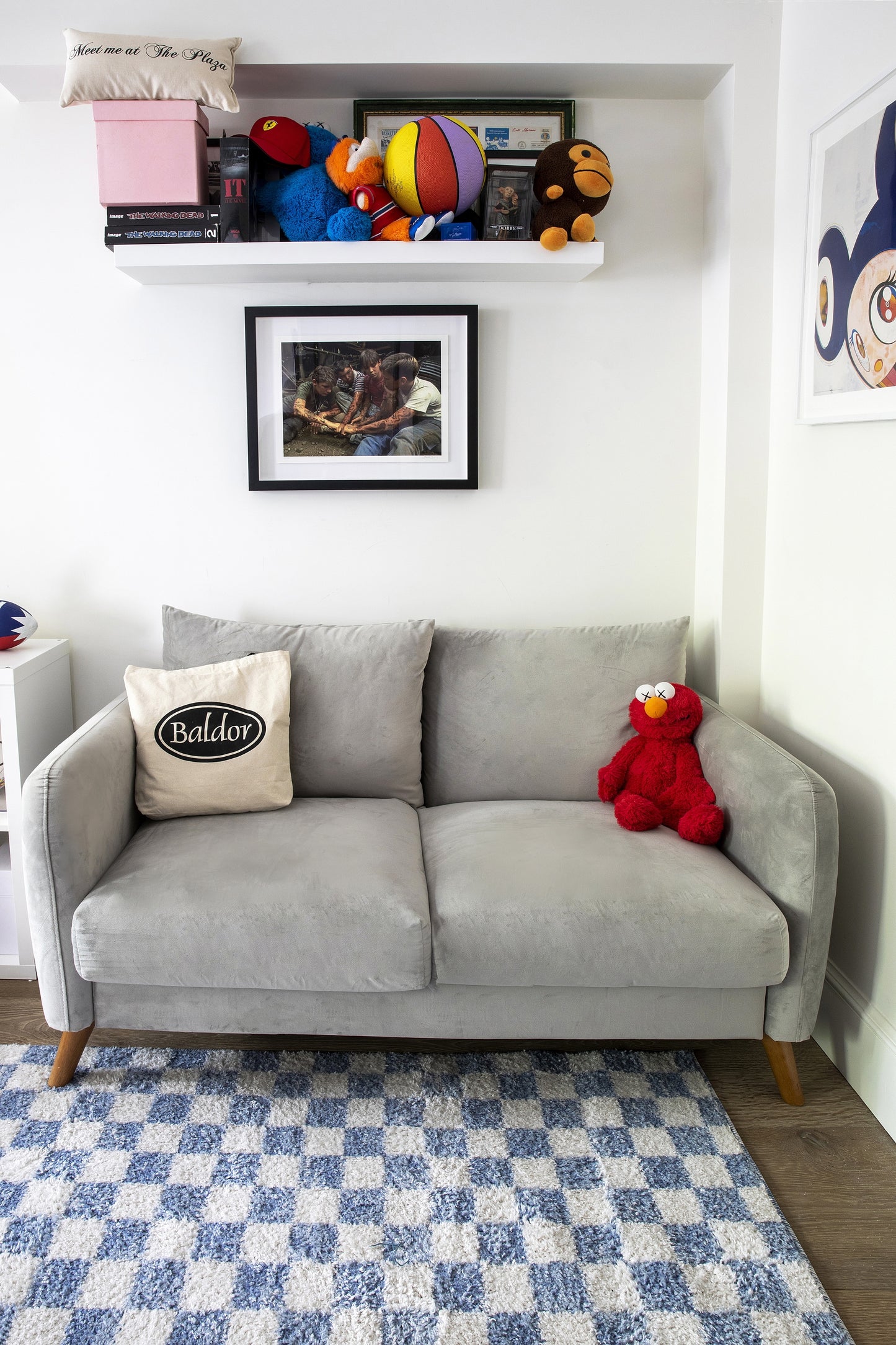 Gray mid-century sofa with wooden legs, red Elmo plush, beige Baldor pillow, blue-and-white checker rug, wall shelf with toys.