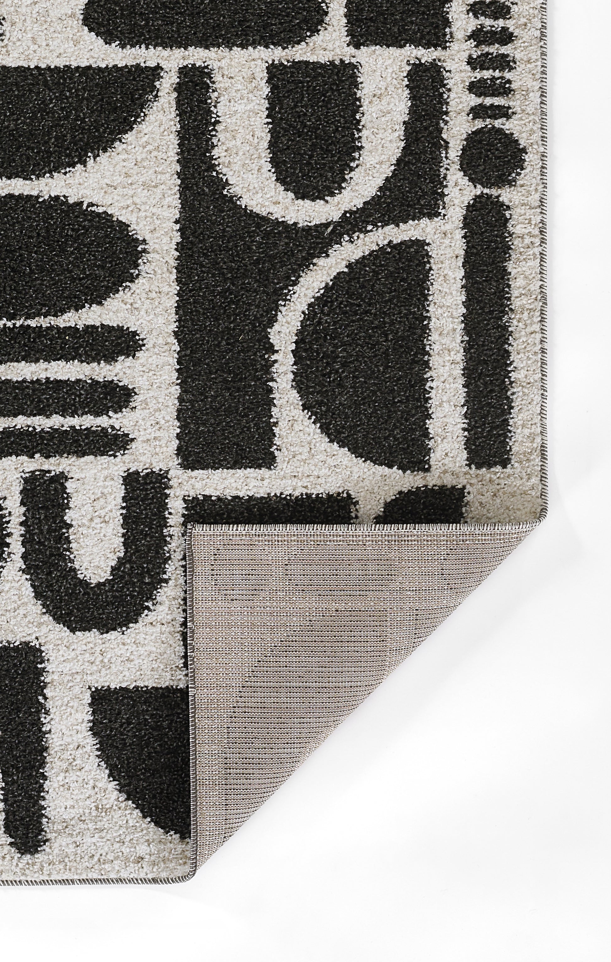 Geometric gray area rug with bold black abstract letter shapes; folded corner reveals beige-brown backing.