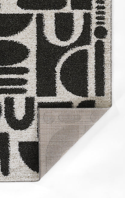 Geometric gray area rug with bold black abstract letter shapes; folded corner reveals beige-brown backing.