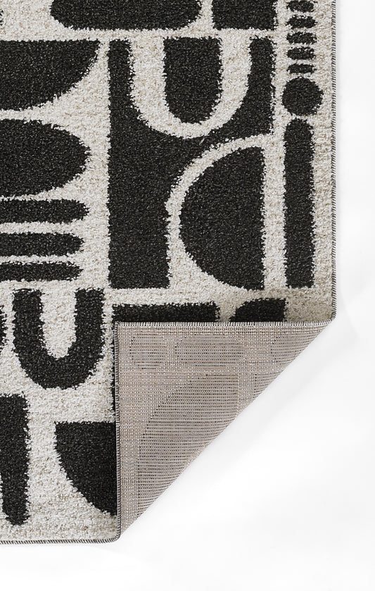 Gray rug with bold black abstract typography motif; folded corner reveals tan non-slip backing.