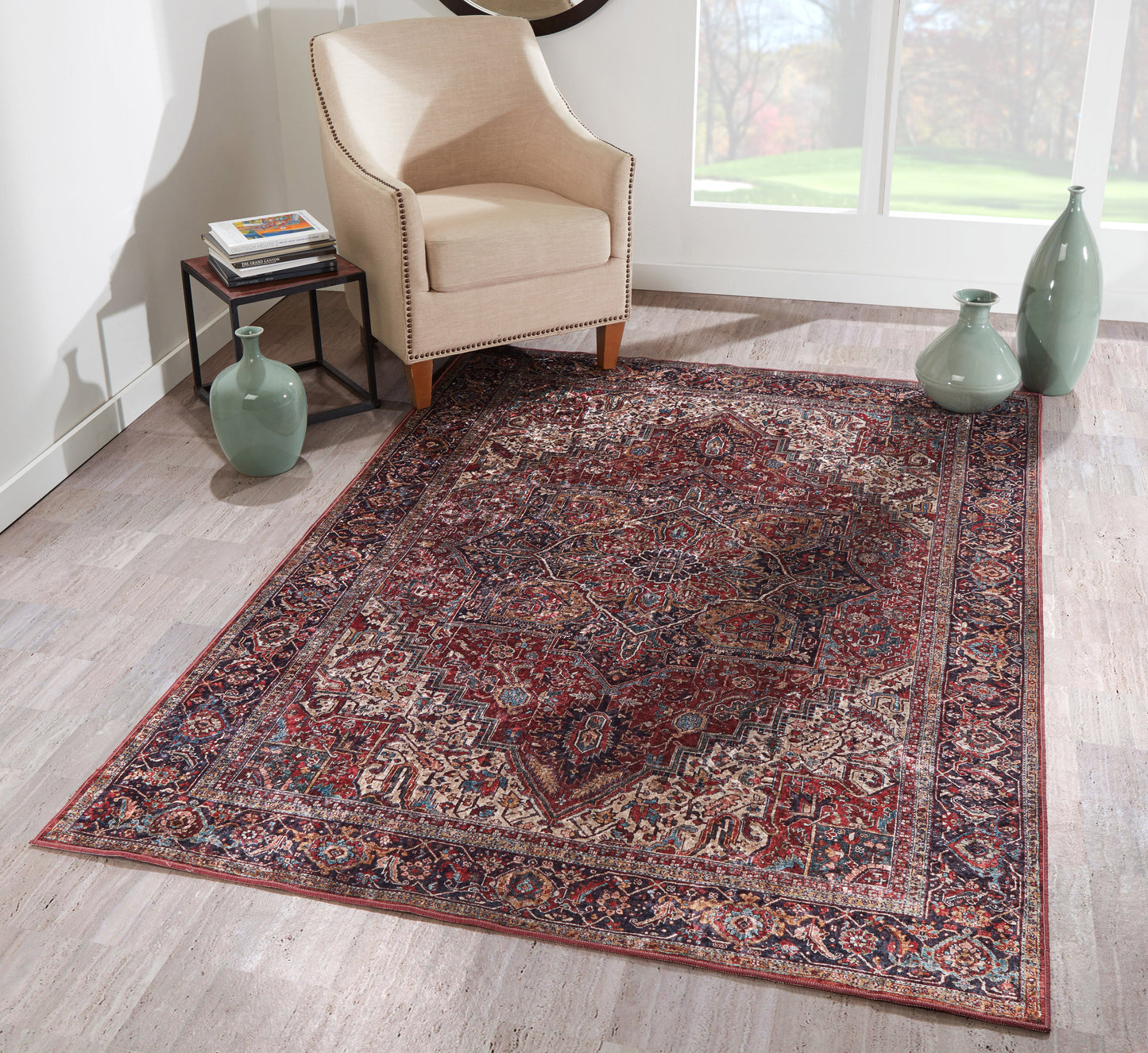Beige armchair with nailhead trim beside a red-and-navy Persian rug on light wood floor; green vases and bright window light.