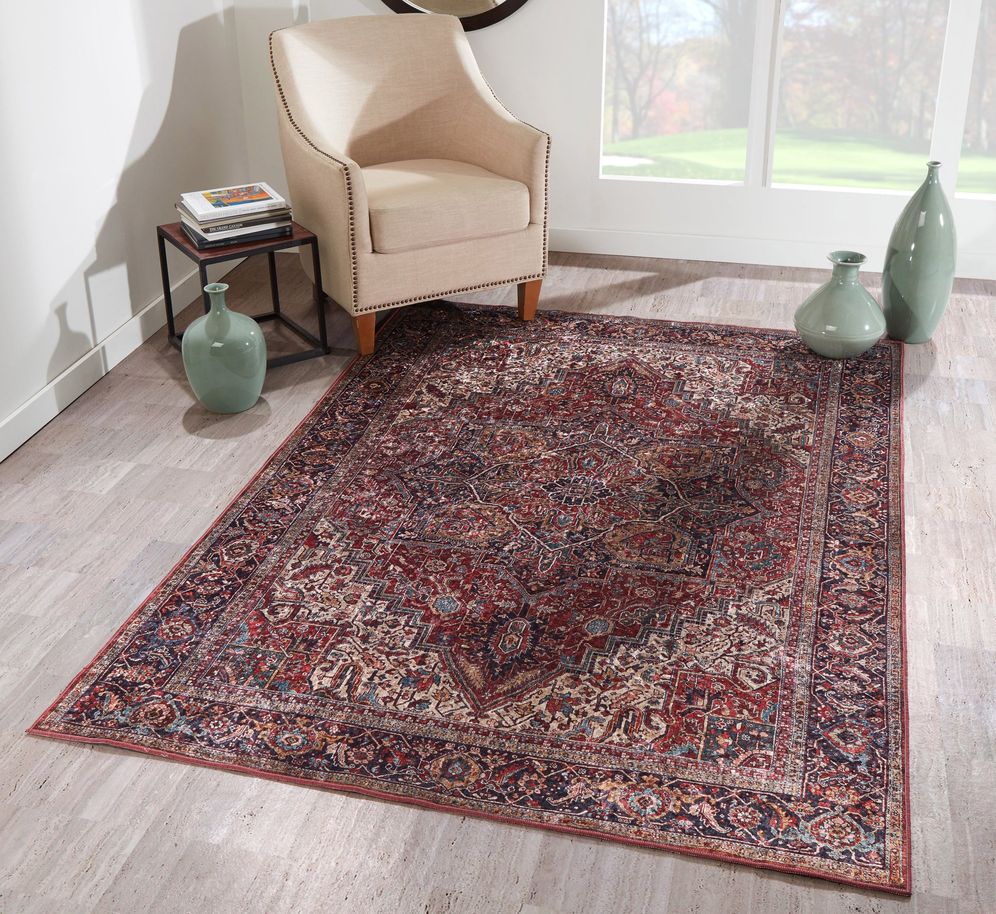 Beige armchair with nailhead trim beside a red-and-navy Persian rug on light wood floor; green vases and bright window light.