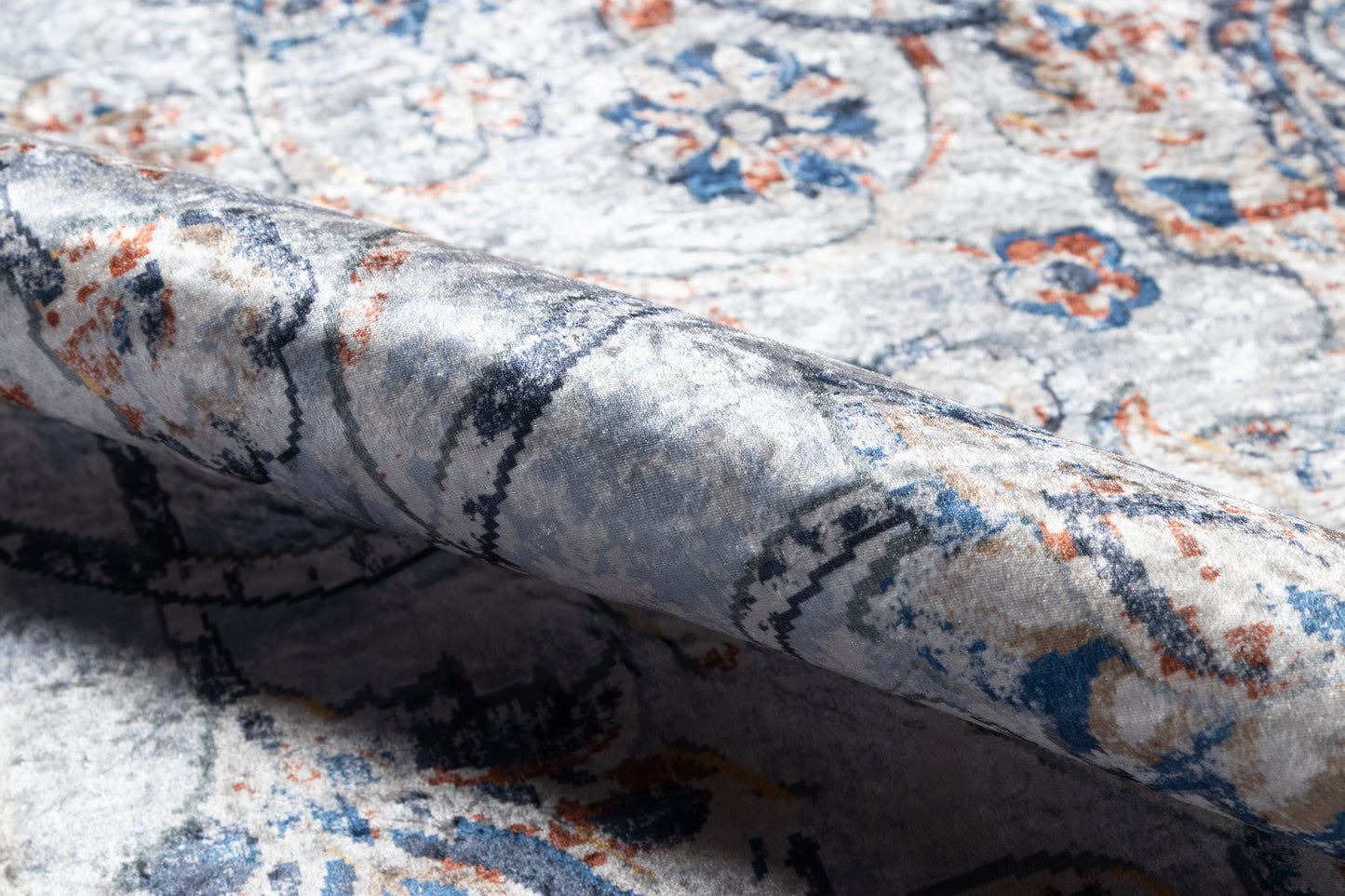 Close-up of an ornate blue-ivory rug with orange accents and floral motifs; folded edge reveals soft, textured pile.