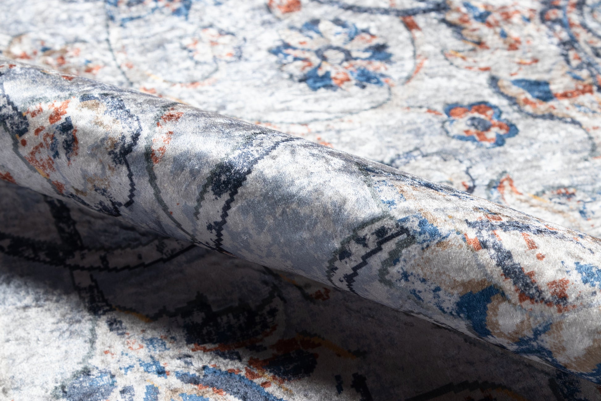 Close-up of an ornate blue-ivory rug with orange accents and floral motifs; folded edge reveals soft, textured pile.