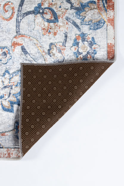 Rectangular area rug with blue and orange floral pattern on light gray; folded corner reveals brown dotted backing.
