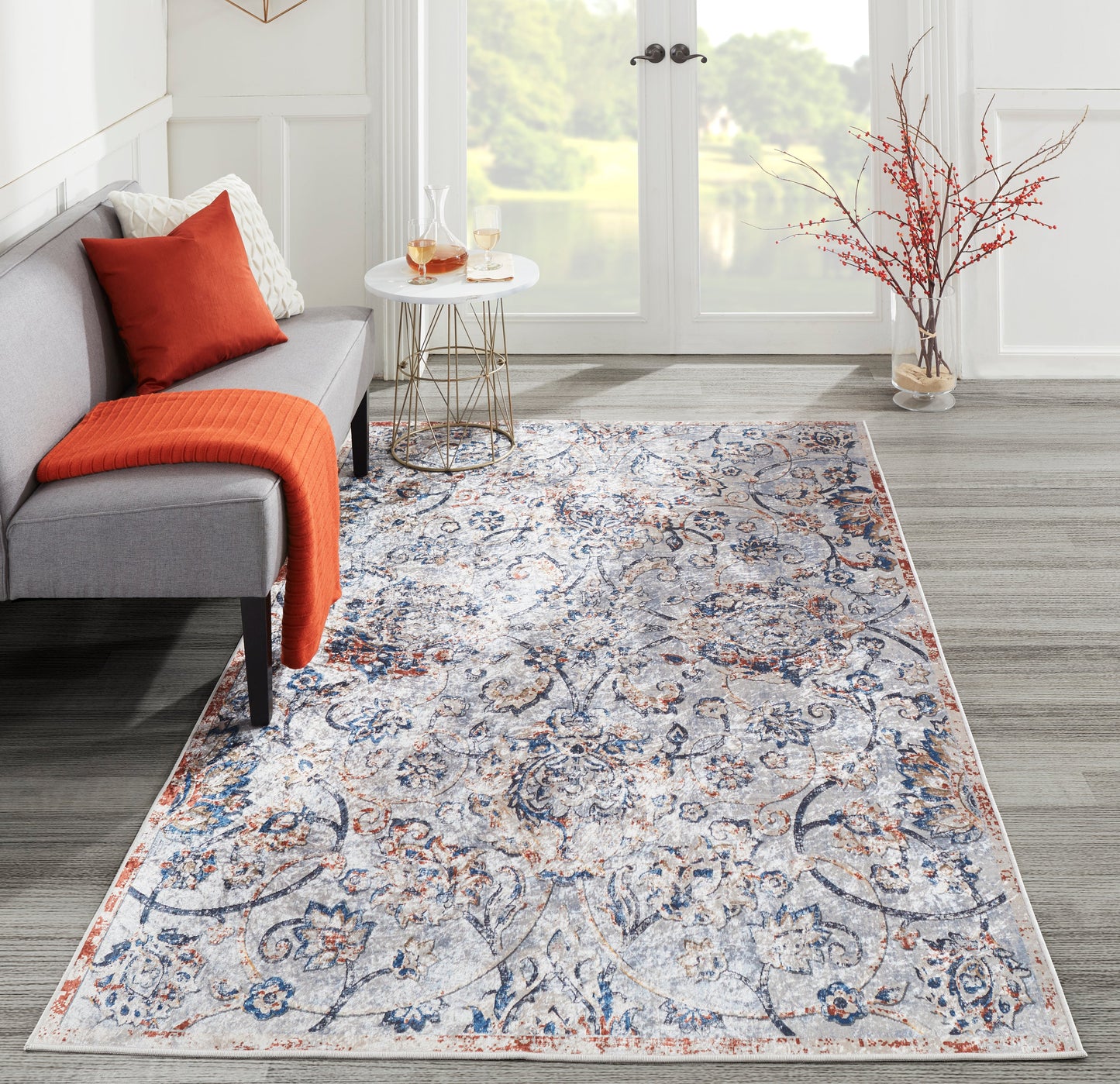 Gray living room with orange throw, blue-and-cream Oriental rug, round glass side table, and vase of branches.