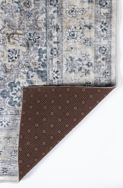 Gray-blue patterned area rug with floral border; folded corner reveals brown dotted underside.