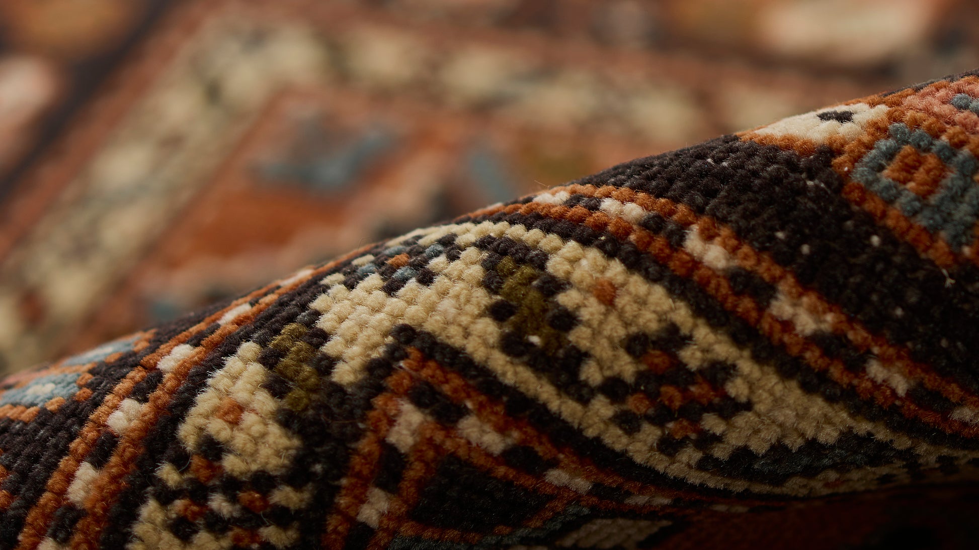 Close-up of a rolled oriental wool rug with bold geometric tribal pattern in cream, brown, blue and orange.