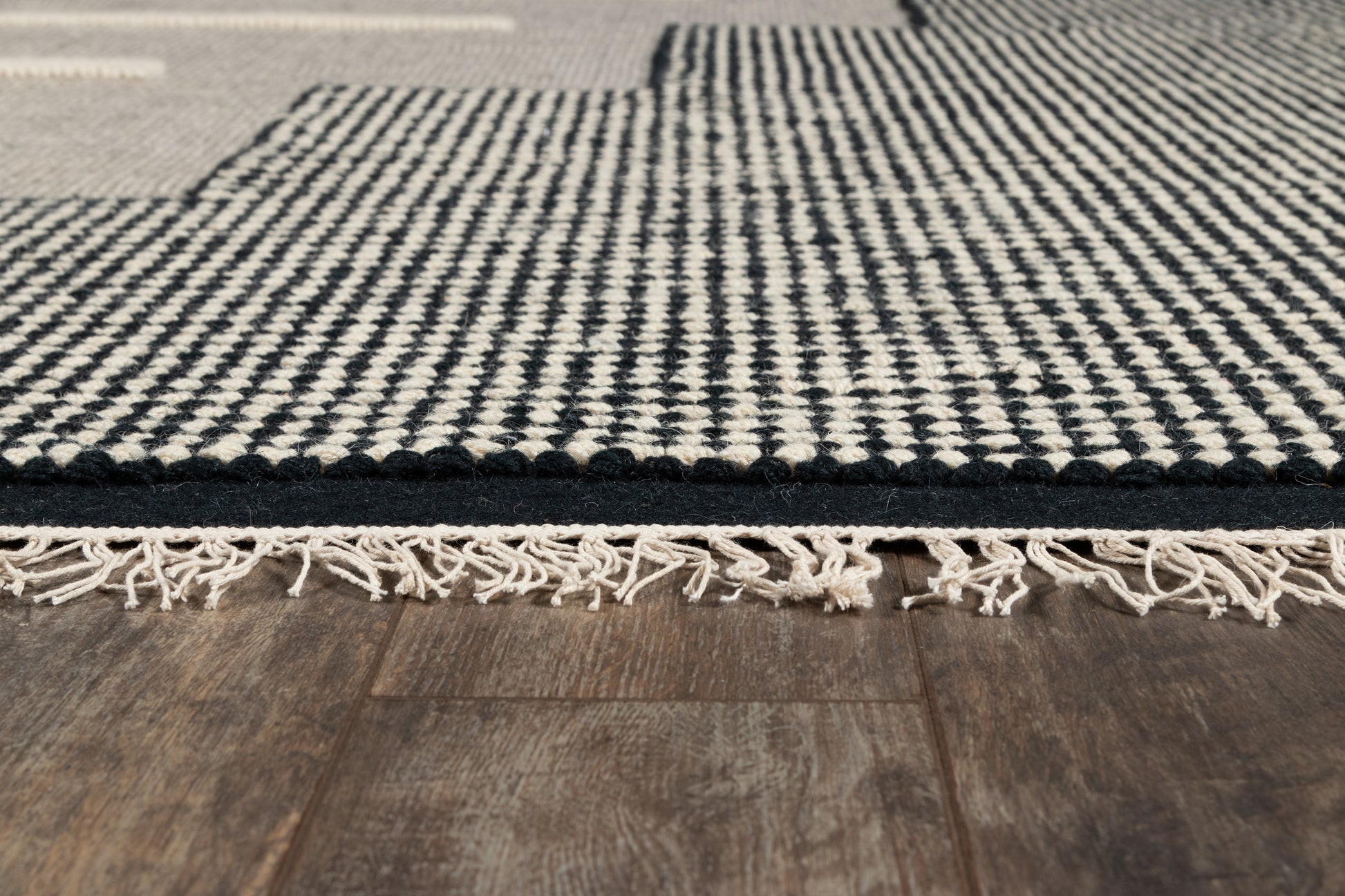 Black and white woven rug with fringe tassels along one edge on dark wood floor.