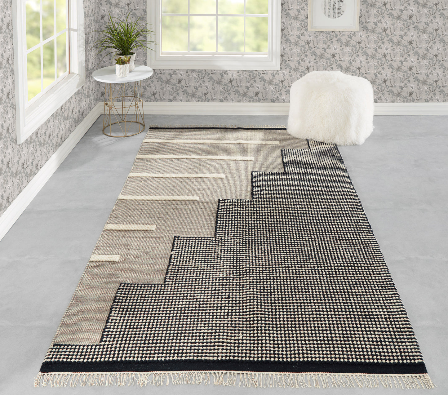 Long woven rug with black and beige stepped zigzag pattern, fringed ends, laid in a bright room with a white fluffy ottoman.