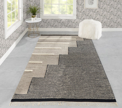 Long woven rug with black and beige stepped zigzag pattern, fringed ends, laid in a bright room with a white fluffy ottoman.