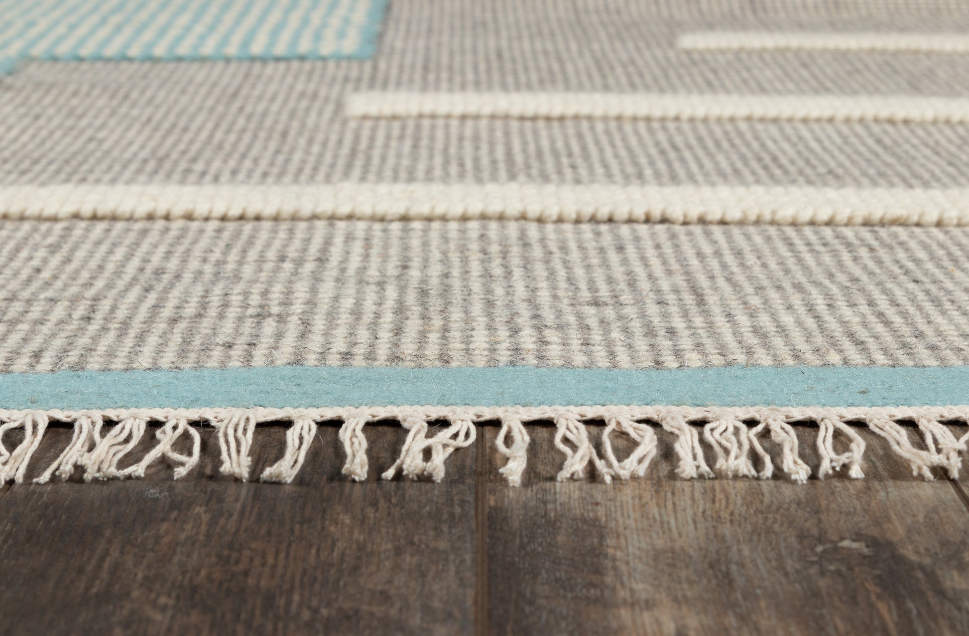 Woven flatweave rug with gray-beige tones, blue stripe, fringed edge on dark wood floor.