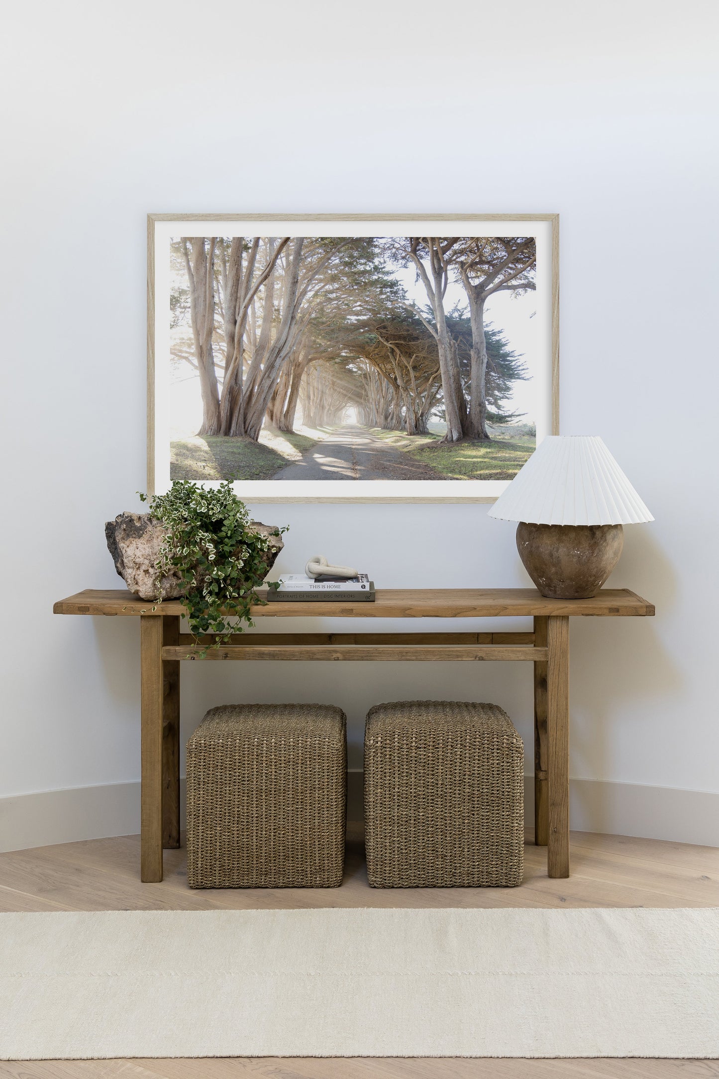 Wooden console table with a framed forest-path photograph, potted greenery, books, a ceramic lamp, and two woven ottomans.