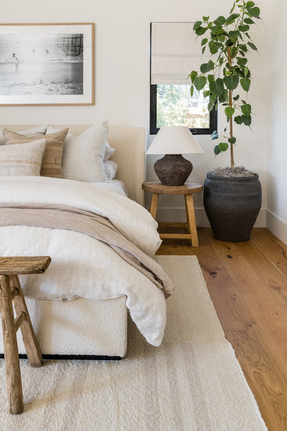 Neutral bedroom with beige upholstered bed, cream linens, wooden stools, a lamp, wall art, and a potted plant by a window.