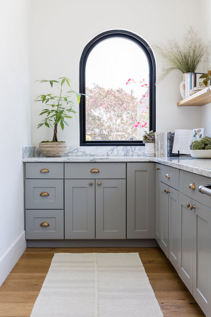 Grey shaker cabinets with brass hardware, marble countertop, arched black window, potted plant, cream rug.