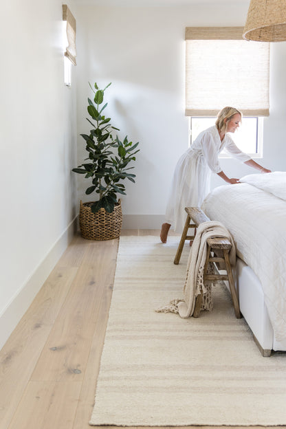 Bright, airy bedroom with white bedding, muted stripe 2'6" x 8' runner on light wood floor, wicker plant basket, woven lampshade.
