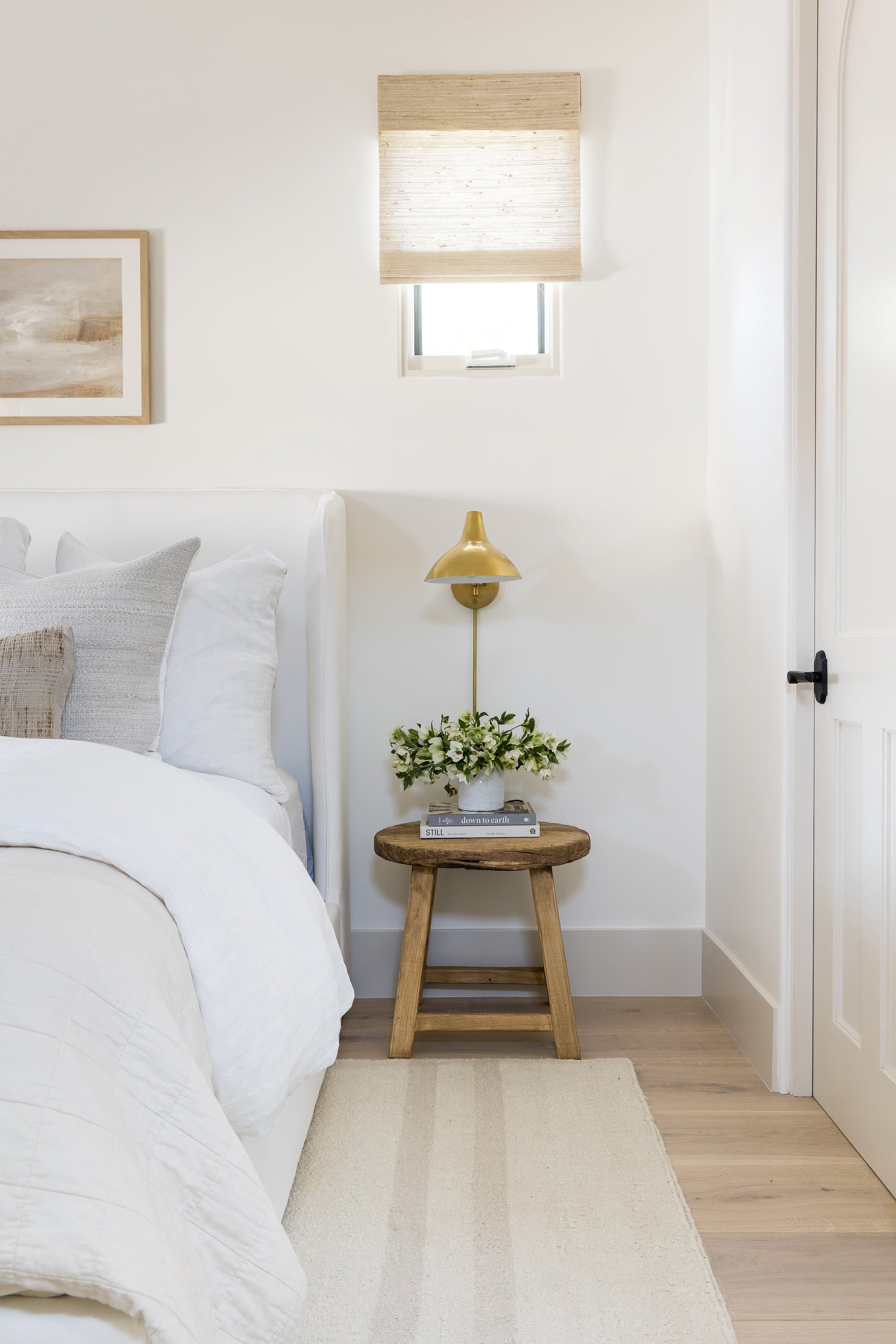 Neutral bedroom corner with white bedding, wooden stool nightstand, brass wall lamp, greenery, books, beige roman shade.