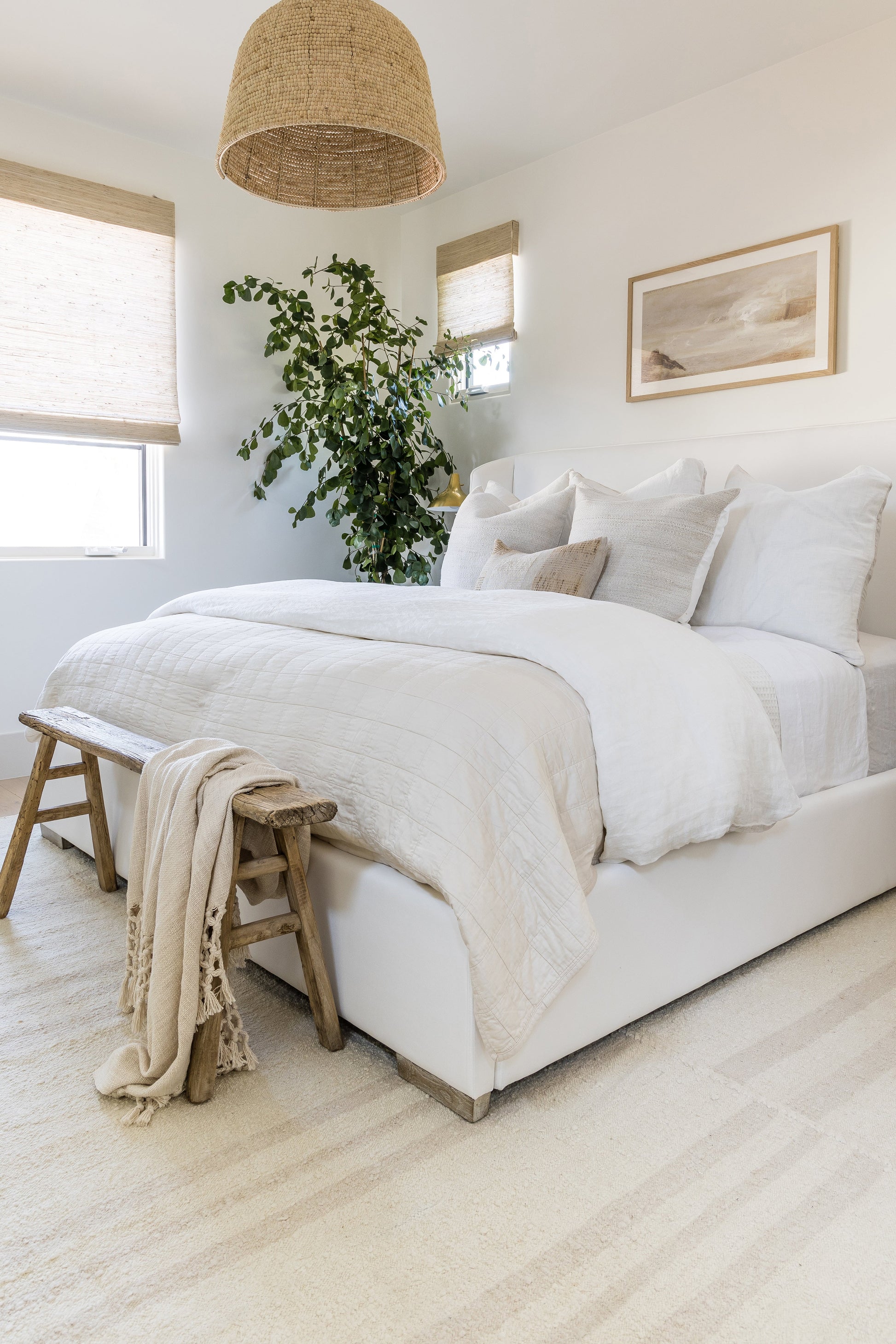 White quilted bedding with beige throw, rustic wooden bench at foot, large indoor plant, woven pendant lamp.