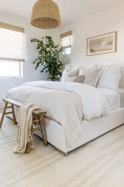 White quilted bedding with beige throw, rustic wooden bench at foot, large indoor plant, woven pendant lamp.