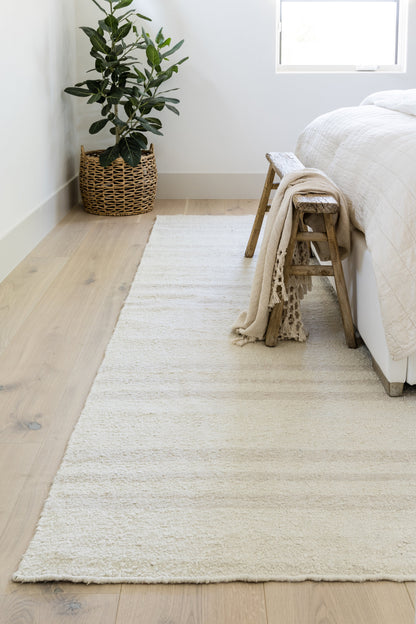 Muted Stripe 10' x 14' rug on light wood floor; cream rug with a muted stripe pattern, beside a bed with a beige throw on a rustic stool and a potted plant in a woven basket.