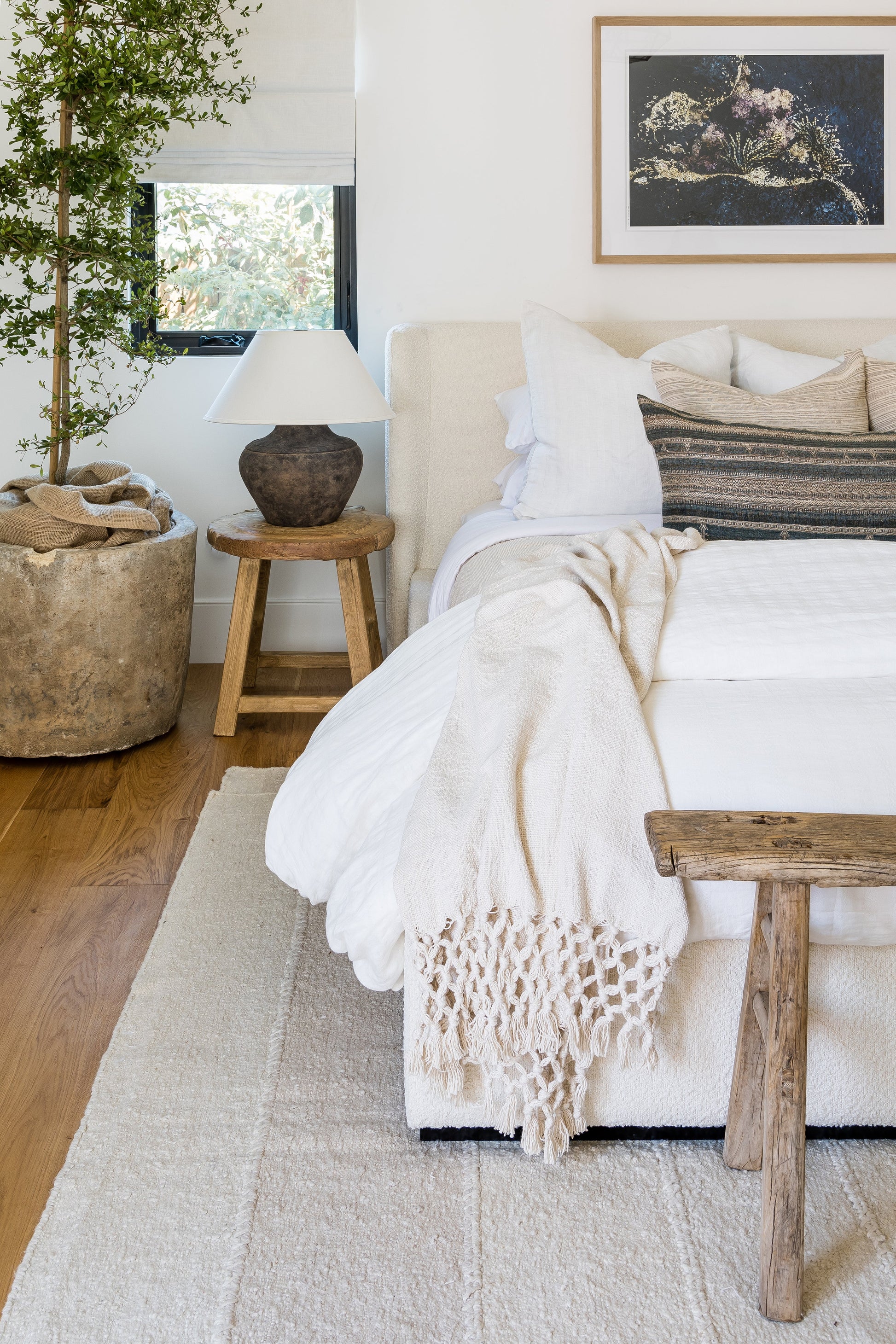 Neutral bedroom with a 2'6" x 8' rug runner on the floor, white bedding, beige fringed throw, rustic wooden bench, potted tree, ceramic lamp, reef art.