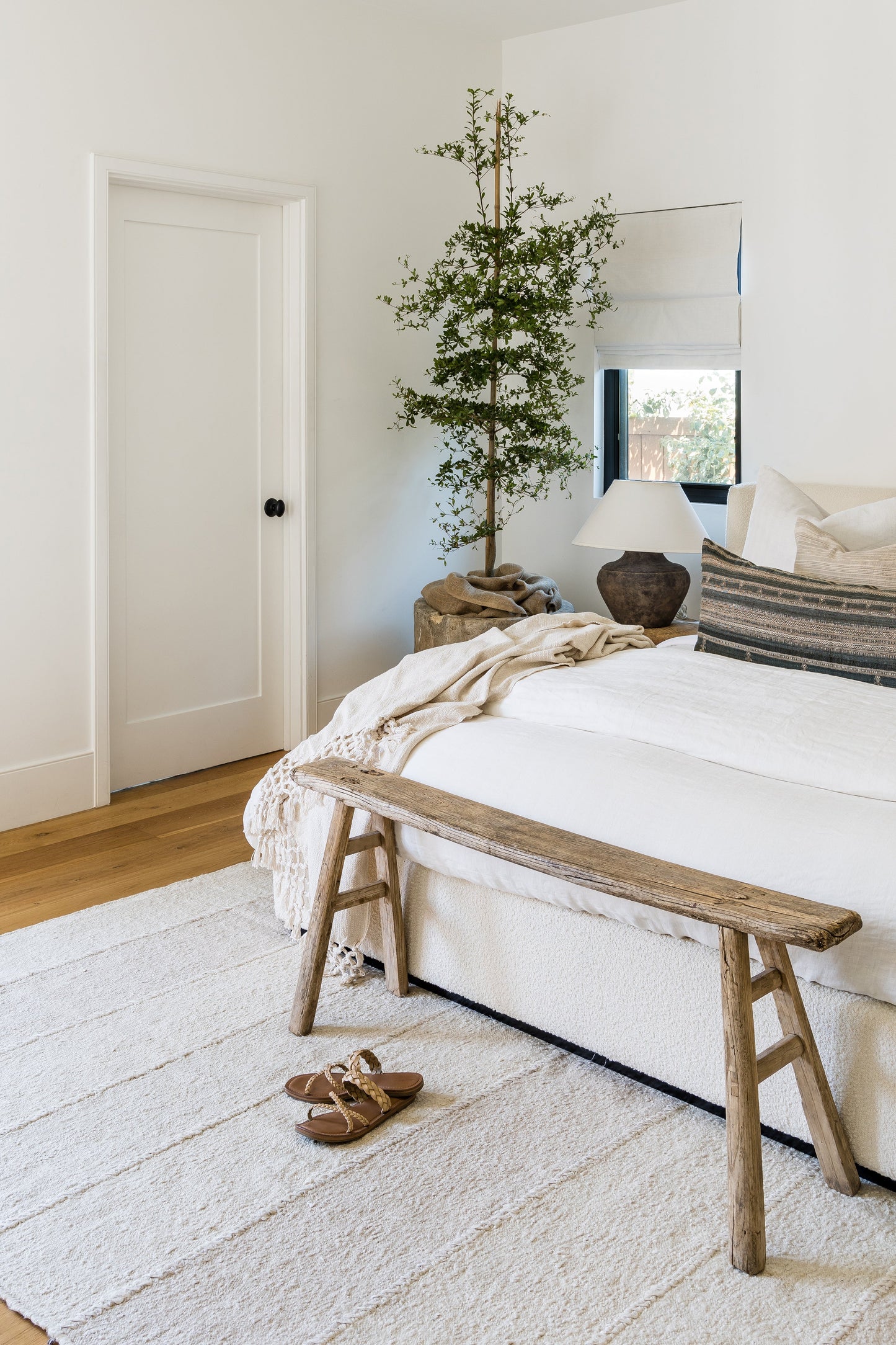 Neutral rug on the bedroom floor in a room with white bedding, a rustic wooden bench at the foot of the bed, a beige throw, striped pillow, tall potted tree, and a ceramic lamp.