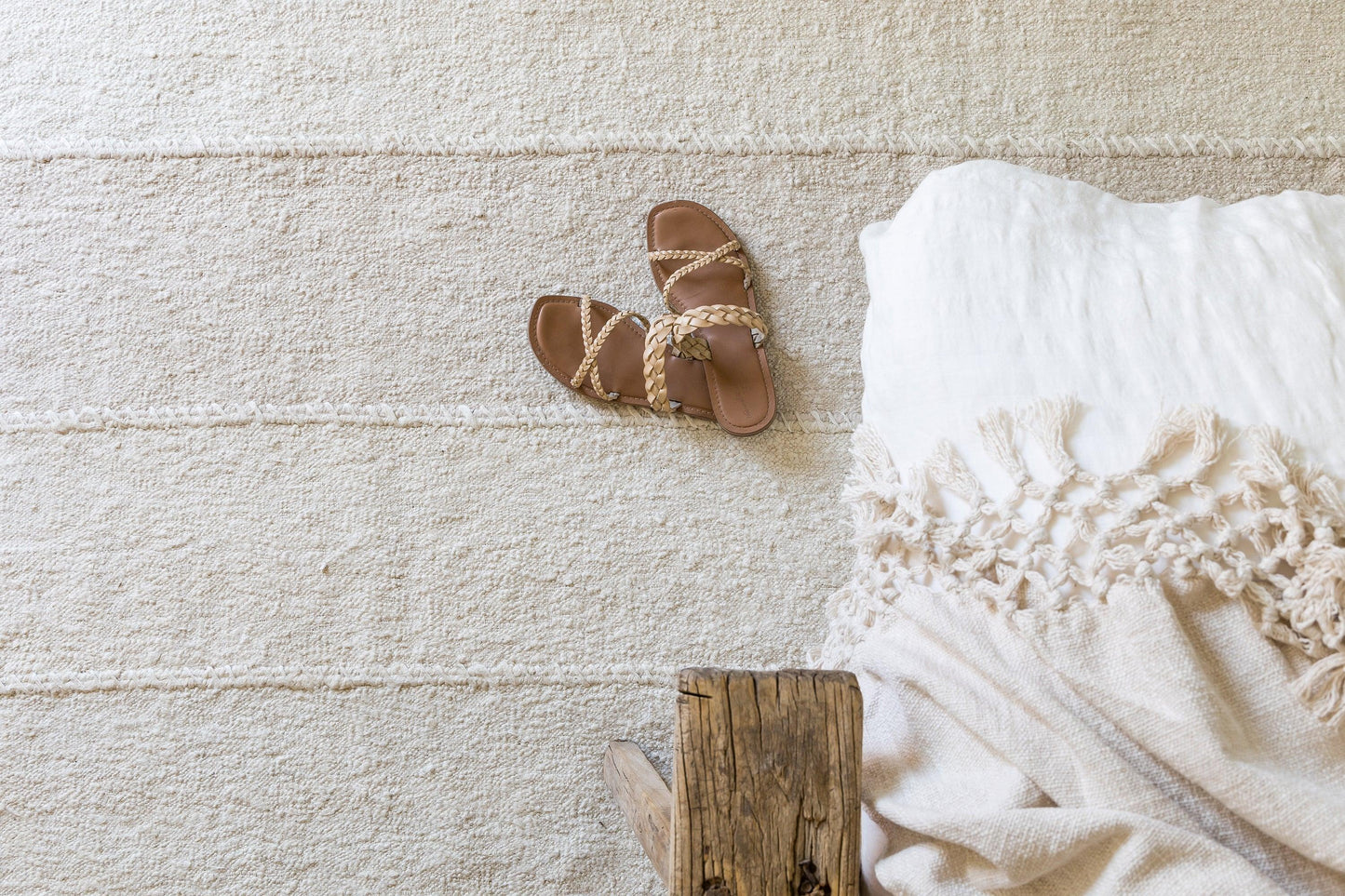 Tan braided-strap sandals on a beige rug beside a white fringed throw blanket and a rustic wooden block.