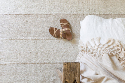 Beige textured rug, brown braided-strap sandals, and cream macrame-fringe throw on a rustic wooden block.