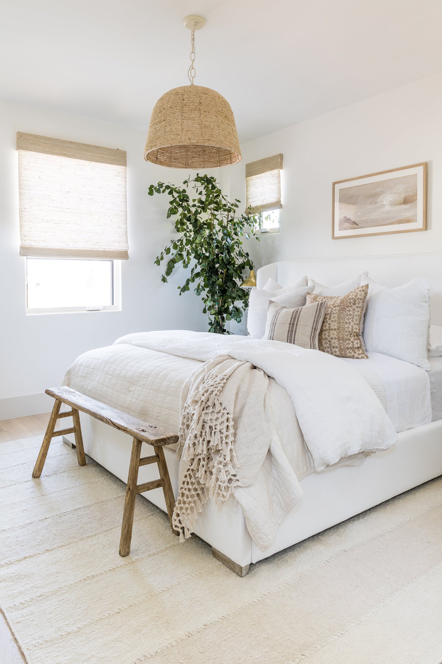 Bright white bedroom with linen bedding, beige throw, wooden bench at foot, woven pendant light, tall plant & neutral art