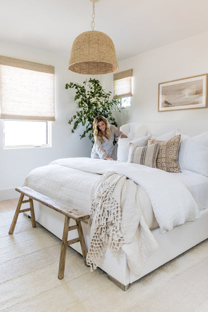 Neutral boho bedroom with rug, white bedding, textured beige throw, rattan pendant lamp, wooden bench, and potted plant.