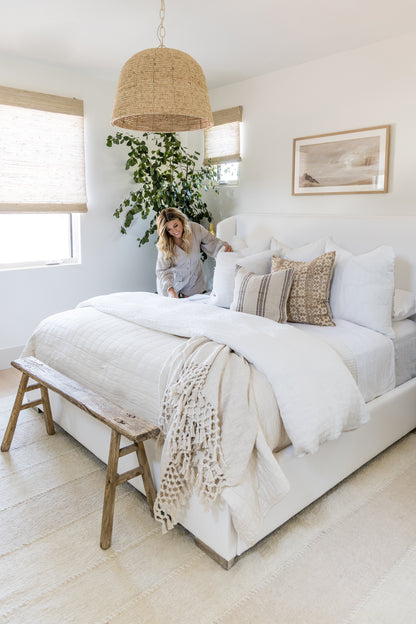 Patchwork rug on the floor of a coastal bedroom; white linen bedding, beige throws, a wooden bench, a woven pendant lamp, and a leafy plant.