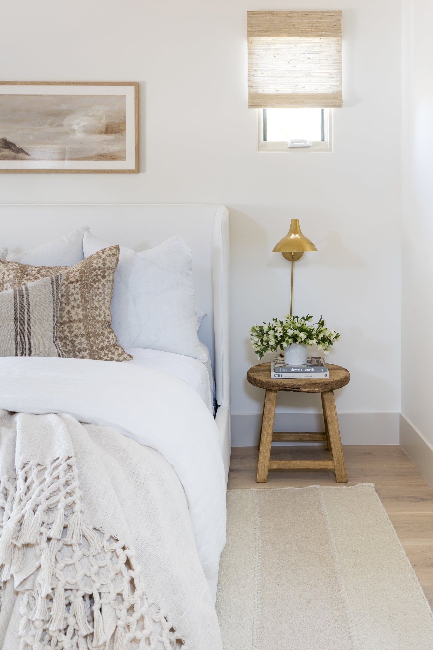 Neutral bedroom scene featuring a 2'6" x 8' rug runner, white linens, beige knit throw, brown patterned pillows, round wooden side table, brass lamp, and greenery.