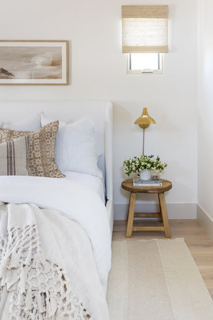 Neutral bedroom with white bedding, woven fringe throw, beige cushions, round wooden nightstand, brass lamp, vase of flowers.