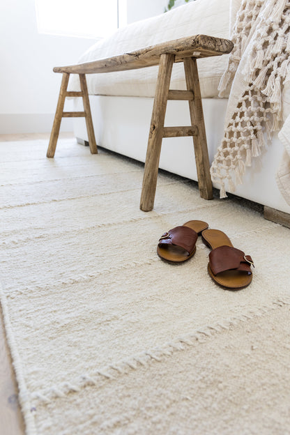 Cream textured rug as the main subject; a rustic weathered wooden bench sits beside a white bed, with brown leather sandals nearby.