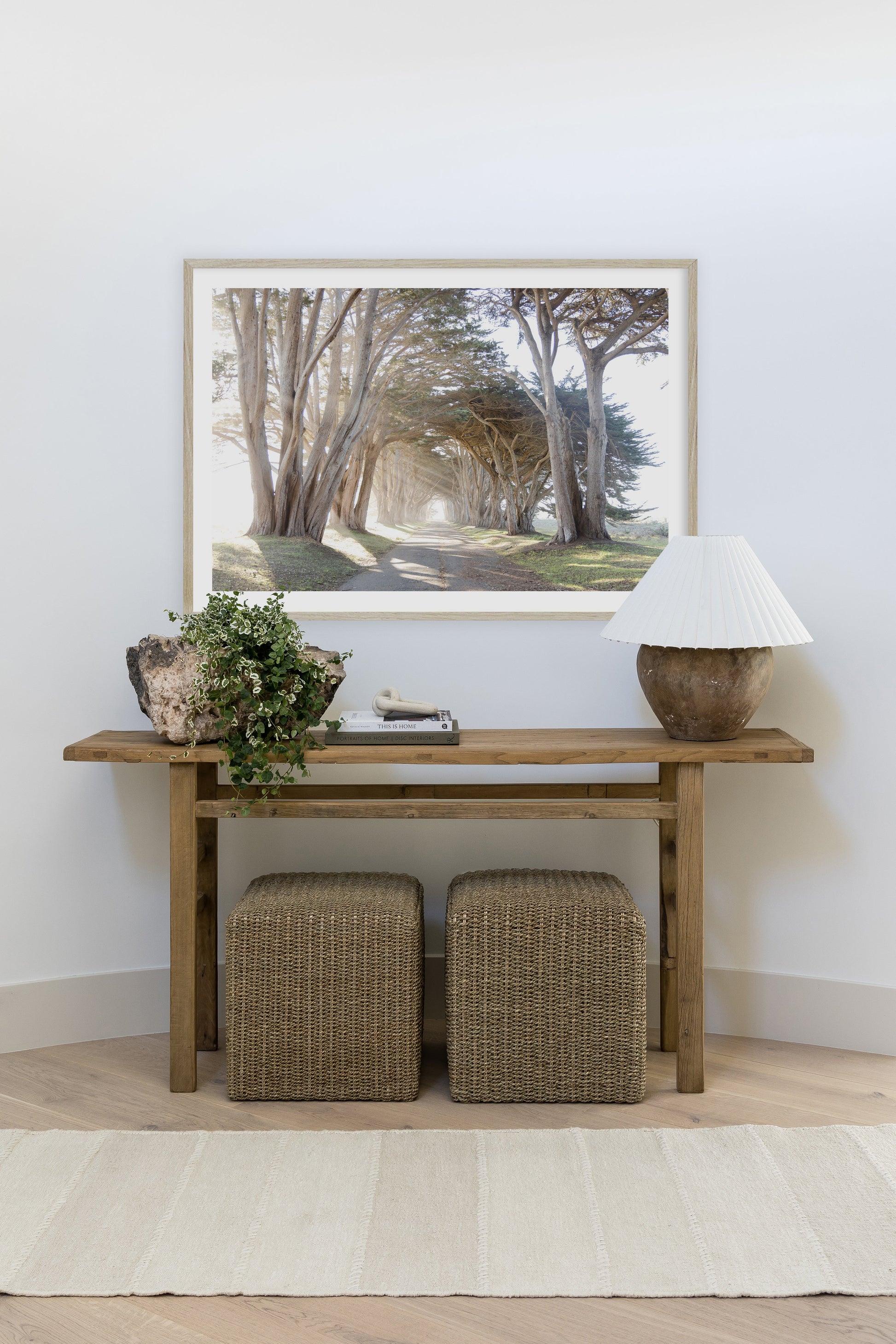 Patch rug 6'3" x 9' in neutral boho decor with a wood console, two woven ottomans, framed path artwork, ceramic lamp and a greenery planter.