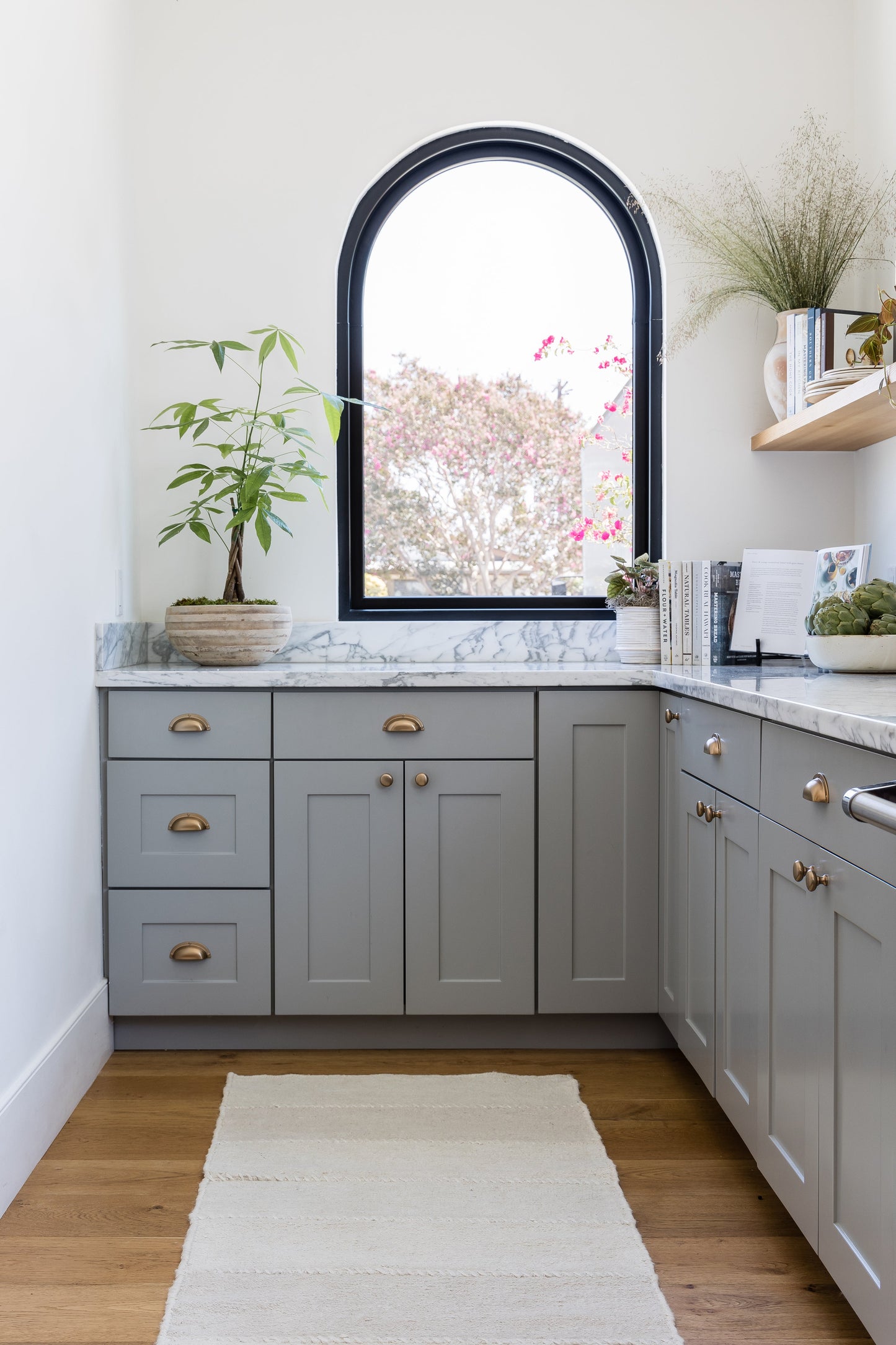 Grey shaker cabinets with brass hardware, marble countertop, arched black window, potted plant, books, cream rug.