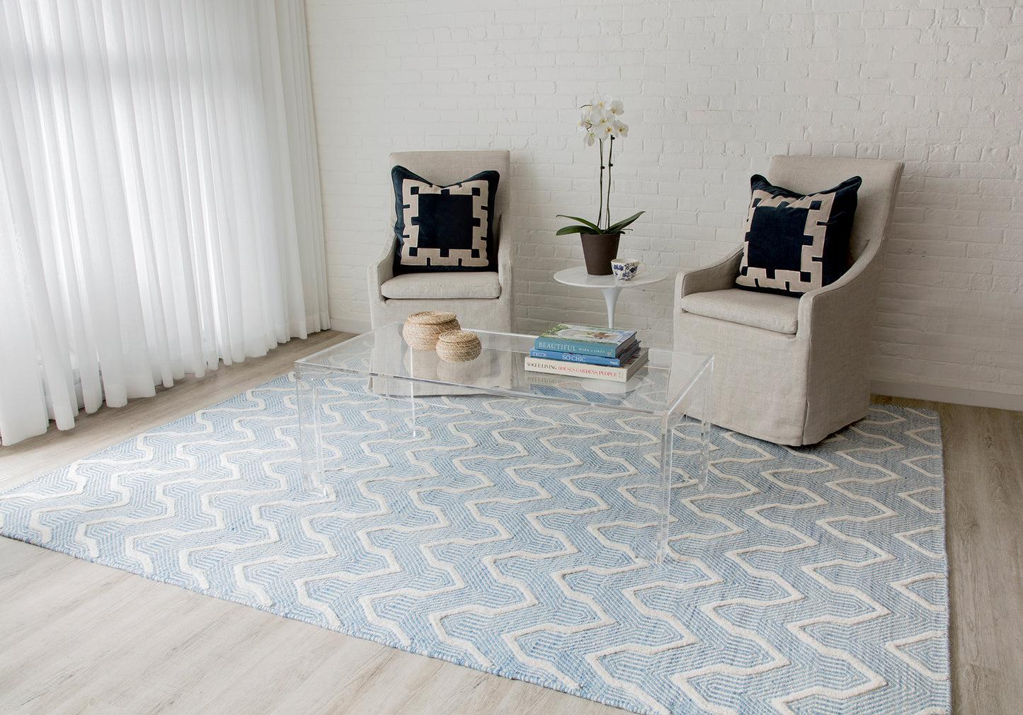 Light blue geometric rug, two beige armchairs with black pillows, clear acrylic coffee table, white brick wall.