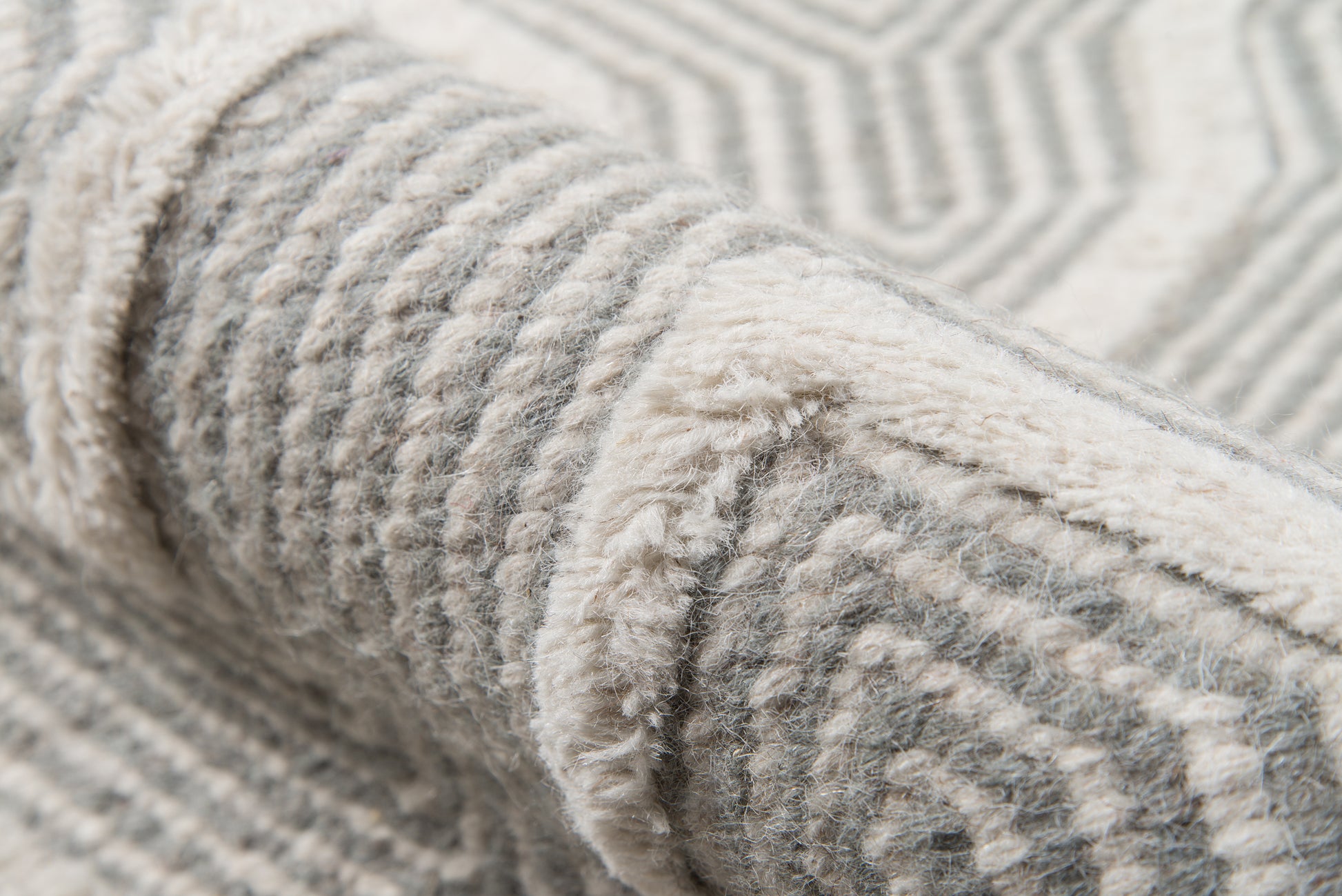 Close-up of a soft gray rug with ivory stripes and geometric pattern, rolled edge showing plush pile.