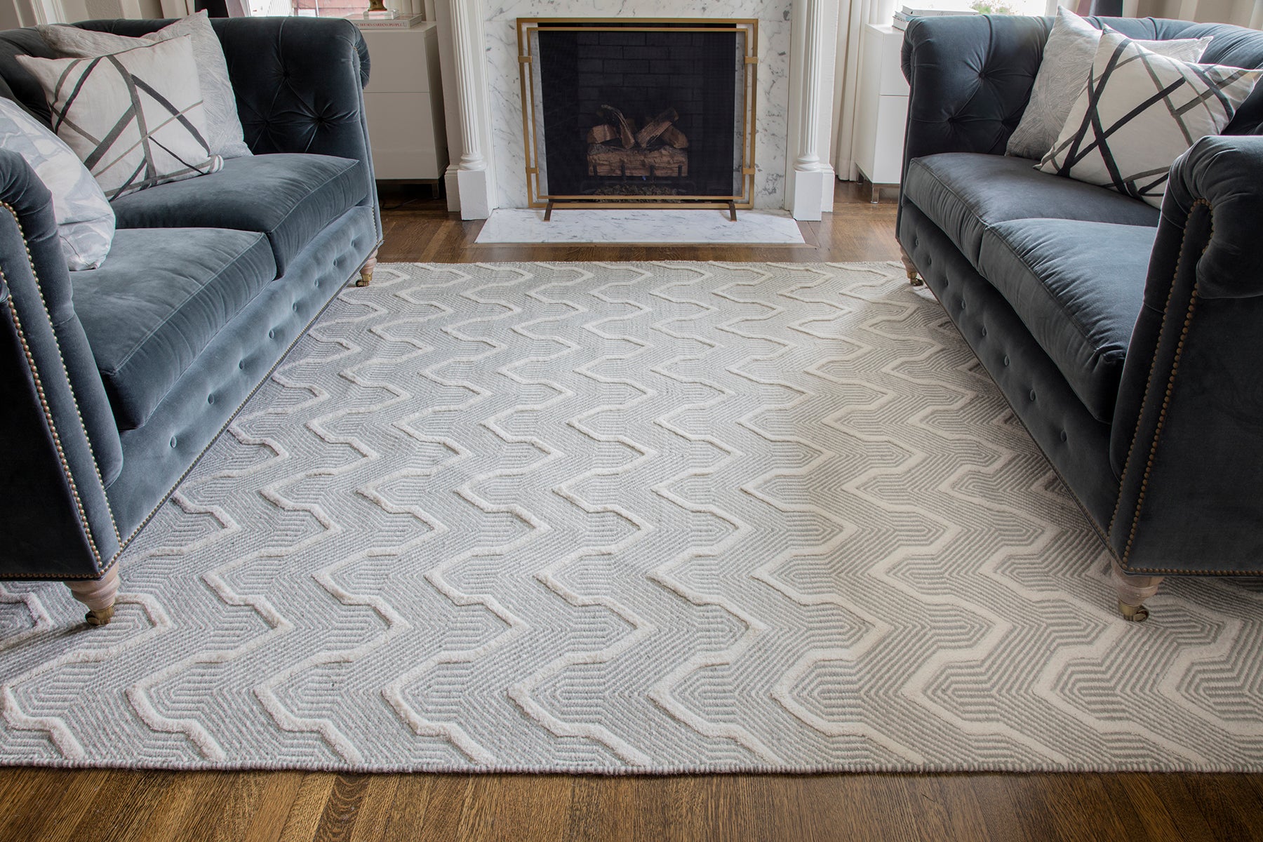 Gray geometric area rug with chevron zigzag pattern between blue-gray tufted sofas in a cozy living room.