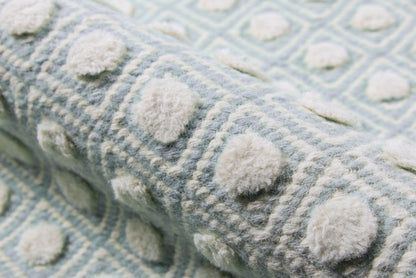Soft blue throw blanket with cream pom-pom bobbles and geometric pattern; cozy wool texture.