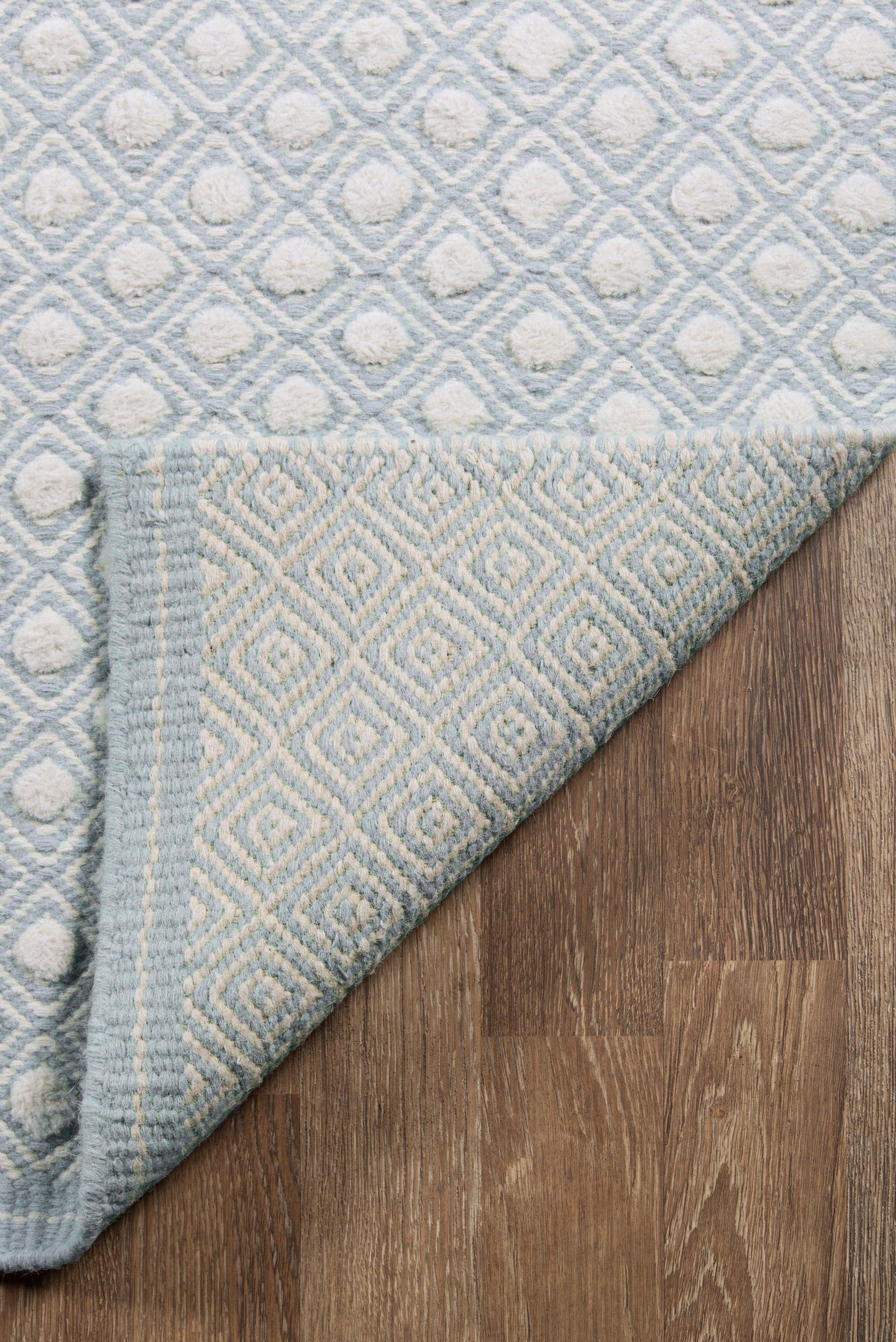 Blue geometric rug with diamond motifs, tufted circles, soft woven texture, folded corner showing underside on wooden floor.
