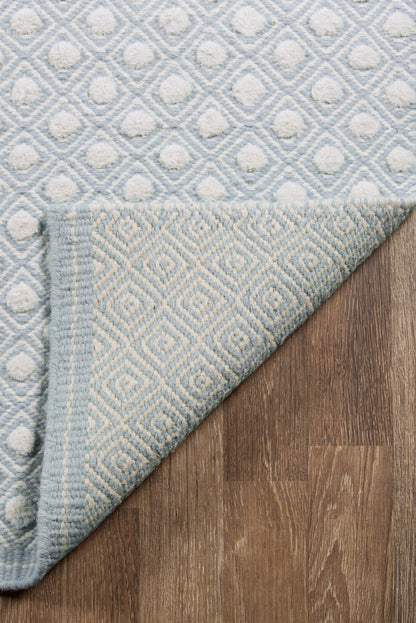 Blue and white geometric rug with diamond lattice; textured tufted surface, folded corner shows underside on wood floor.