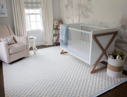White baby crib with wooden X-frame legs, blue blanket, in a gray-blue nursery with a beige armchair and patterned rug.