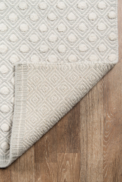 Grey geometric diamond patterned rug with soft cream raised motifs, rolled corner showing underside, on wood floor.