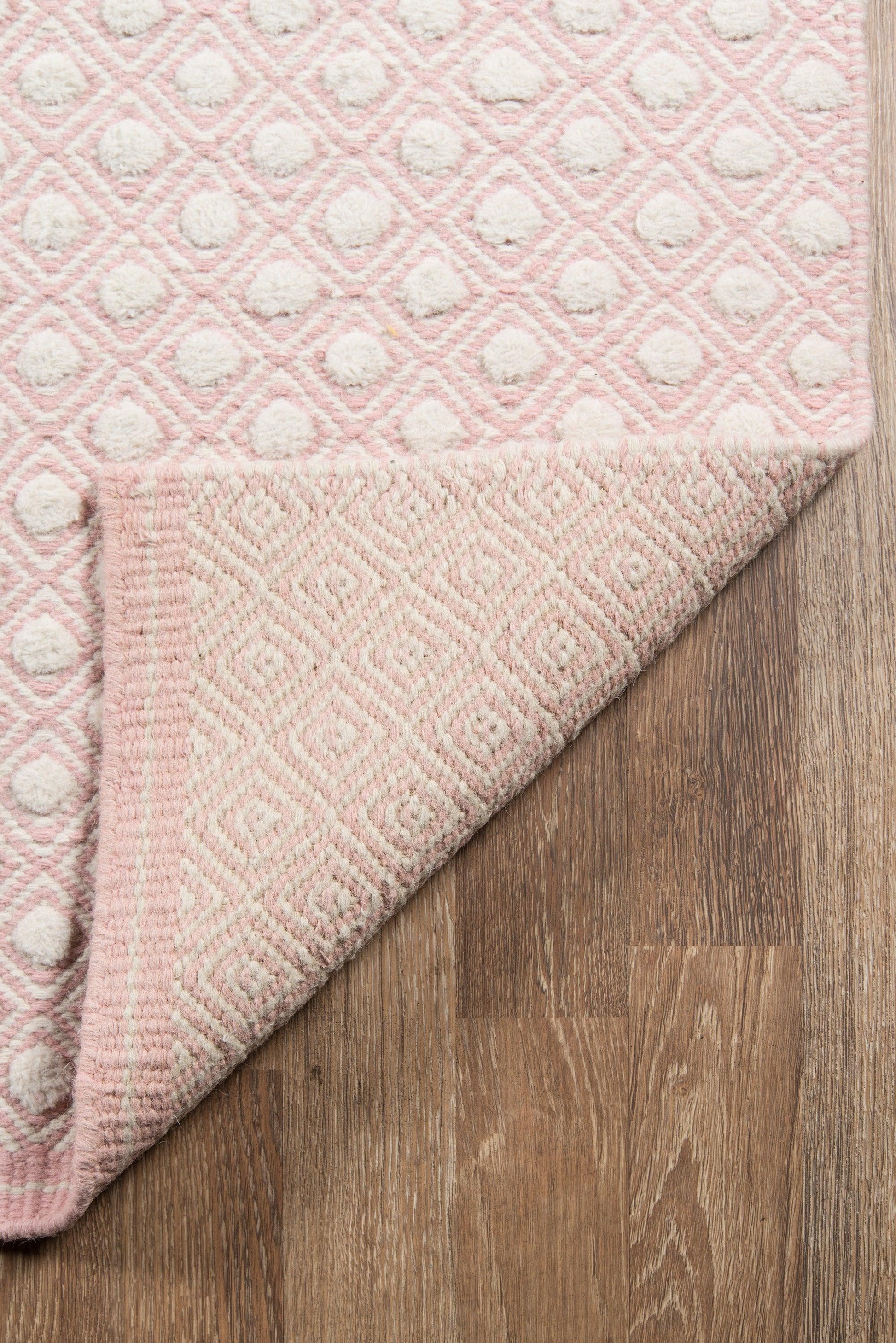 Pink and ivory geometric tufted rug with diamond pattern and raised dots, folded corner shows underside on wood floor.