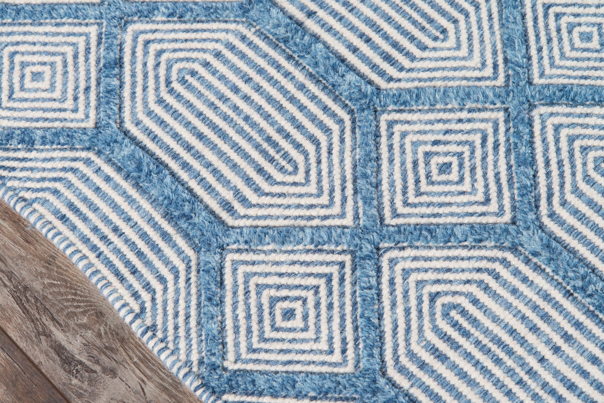 Blue geometric rug with white concentric square spiral patterns, textured pile, corner against a wooden floor.