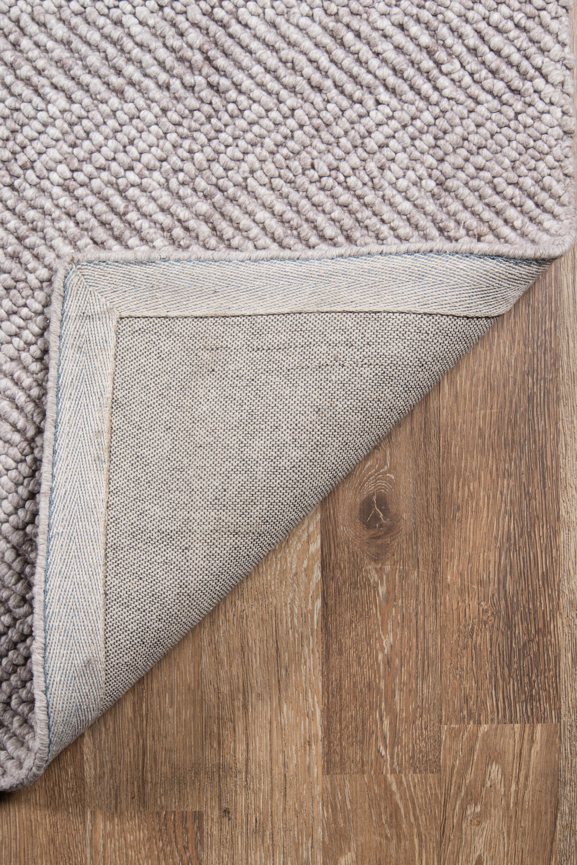 Beige-brown loop-pile rug with folded corner showing grey backing and stitched edge on wooden floor.