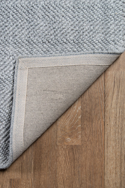 Grey textured loop-pile rug with folded corner exposing the underside on brown wood floor.