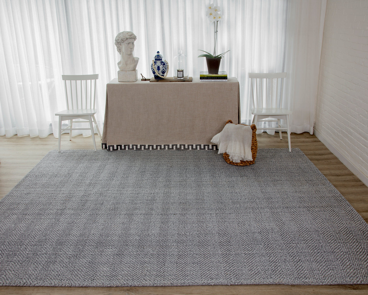 Large rectangular gray textured rug on light wood floor in a bright minimalist room with white chairs and beige table.